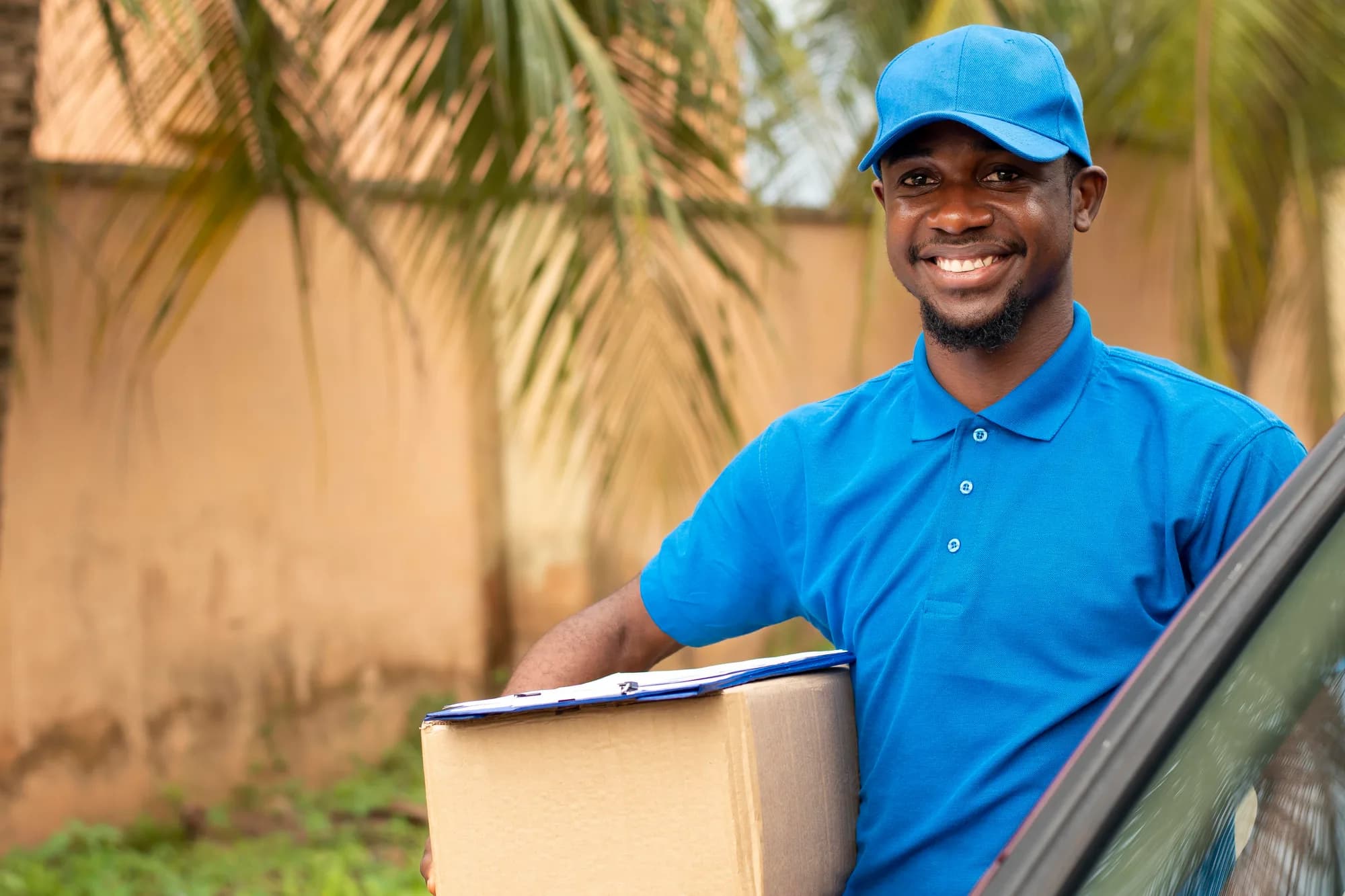 Un livreur souriant en polo bleu tient un colis devant un mur ocre et des palmiers.