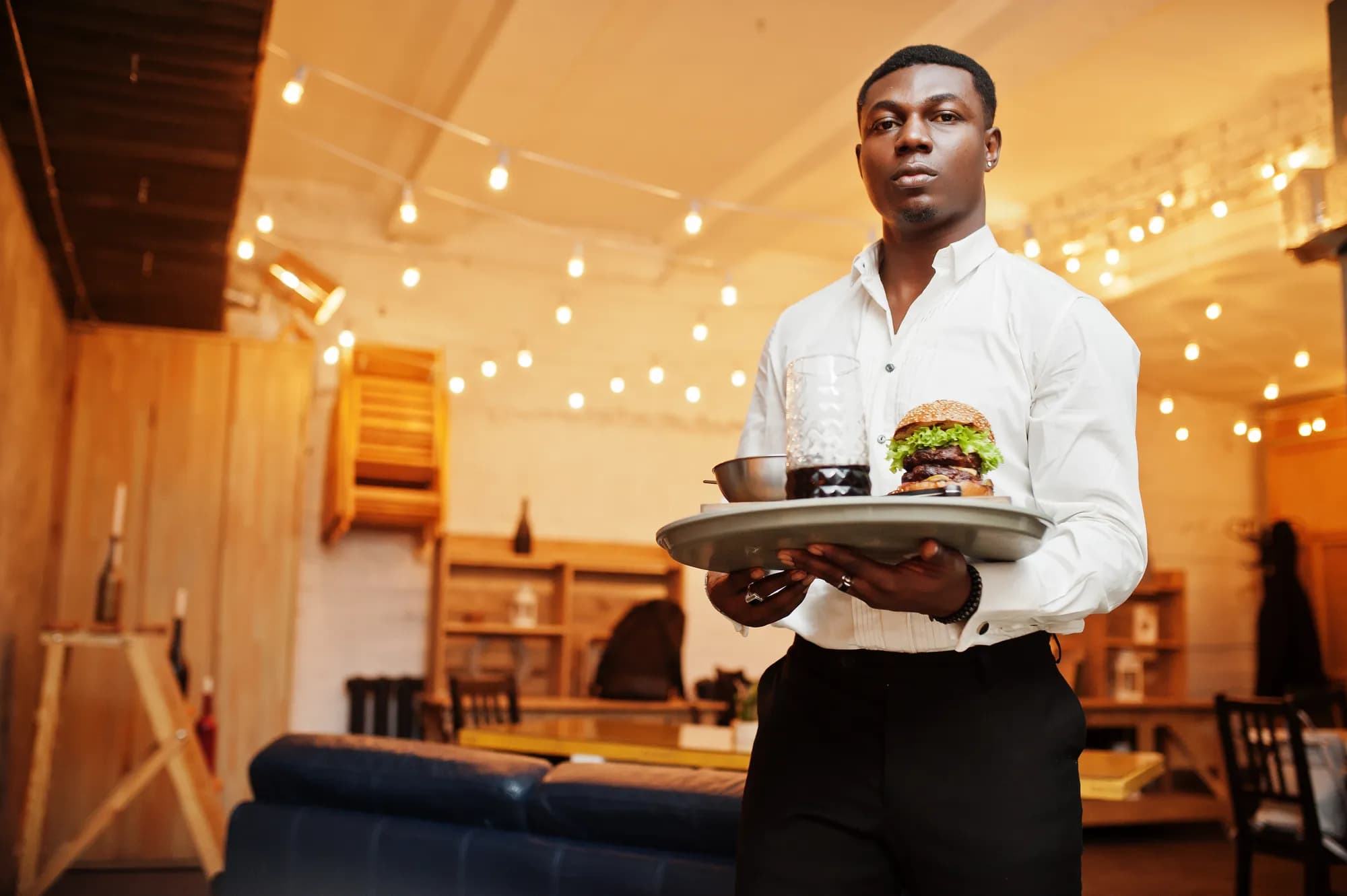 Un serveur en chemise blanche apporte un plateau dans un restaurant décoré de guirlandes.